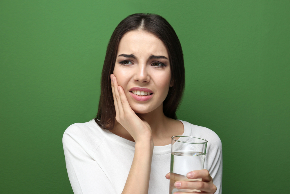 young woman experiencing pain after dental visit, hand up to her cheek, holding a glass of water, on green background