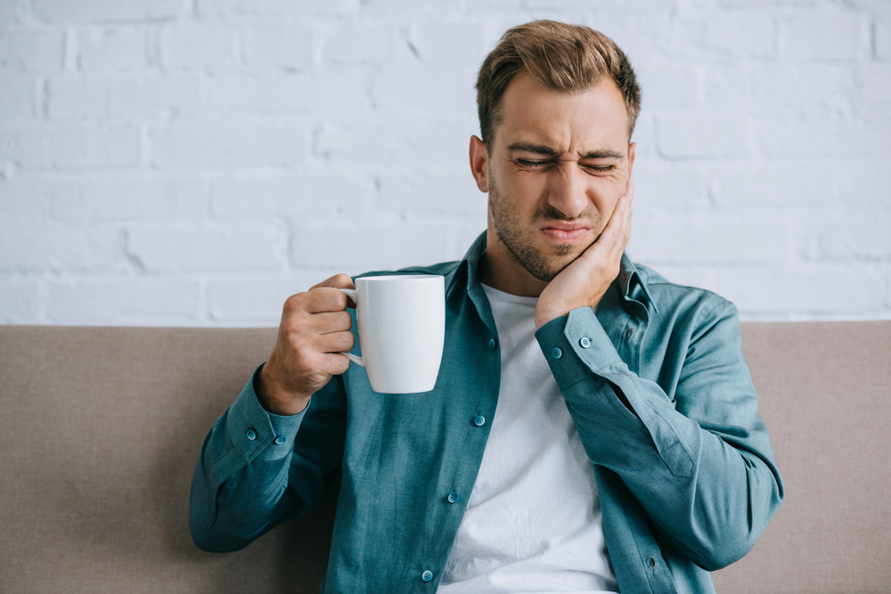 man wincing because of TMJ pain holding coffee mug, sitting on couch