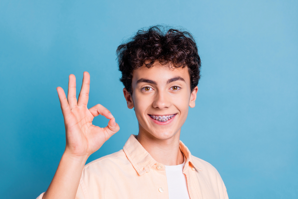 happy young man with curly hair and braces holding up an "ok" sign with fingers on blue background