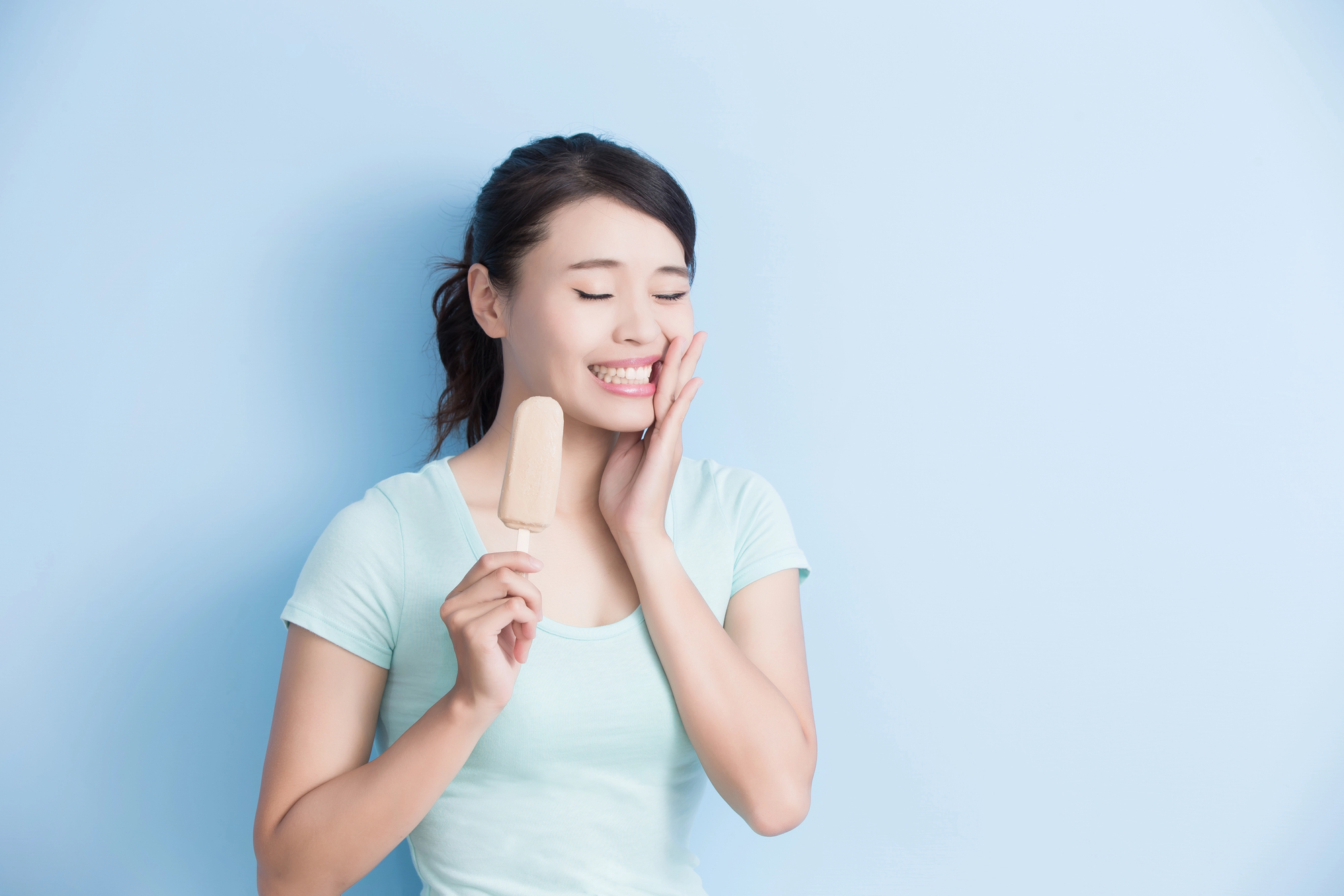Woman smiling with eyes closed, holding a beige popsicle near her face against a pale blue background.