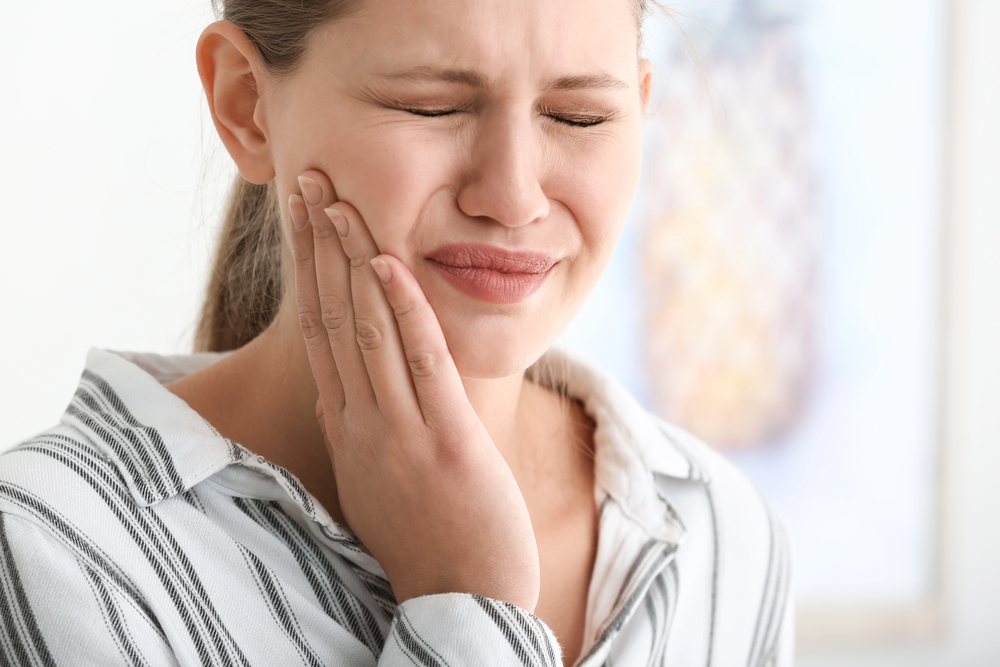 Woman touching her cheek with a pained expression, eyes closed, as if suffering from toothache or facial pain on a striped shirt backdrop.