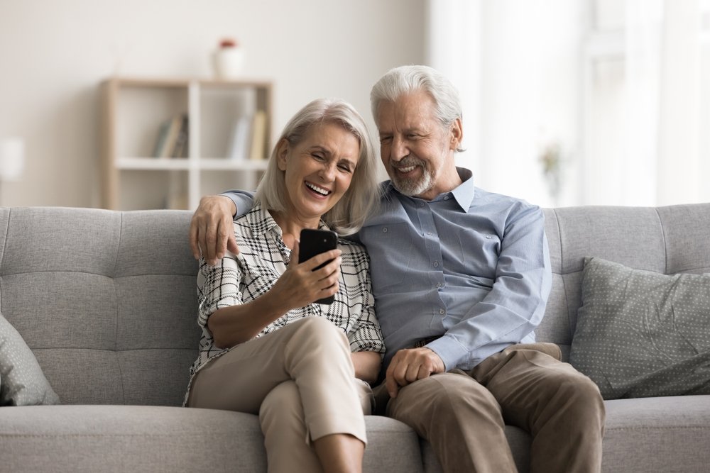 HappyAgedWifeAndHusbandSitOnSofaLookAt Older couple reviewing Medicare dental coverage options at home